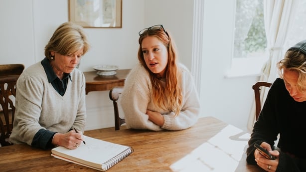 Two younger woman sit around a dining table with an older woman who takes notes on a notepad.